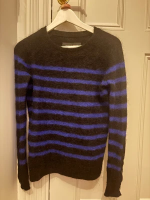 Randig svart-blå stickad tröja By Malene Birger - Mysig stickad tröja från By Malene Birger i svart med blå ränder. Tröjan har lång ärm med knappar på ärmsluten och en rund halsringning. Materialet är fluffigt och ger en cool look, perfekt för dig som gillar att sticka ut med mönster.