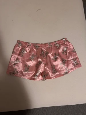 Rosa satinshorts med kaninmönster - Säljer ett par söta rosa shorts i satin med gulligt kaninmönster över hela tyget. De har en loose passform och resår i midjan för extra komfort. Perfekta för chill hemma eller som pyjamasshorts. Supermjuka och glansiga!