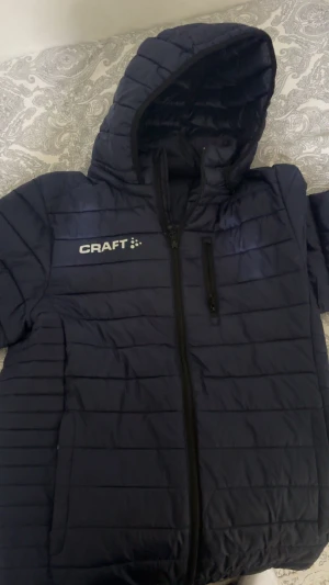 Mörkblå pufferjacka från Craft - Säljer en mörkblå pufferjacka från Craft i storlek S. Jackan har huva, dragkedja framtill och en bröstficka med dragkedja. Quiltad design med horisontella sömmar och logga på bröstet och nedre ryggen. Perfekt för kalla dagar.