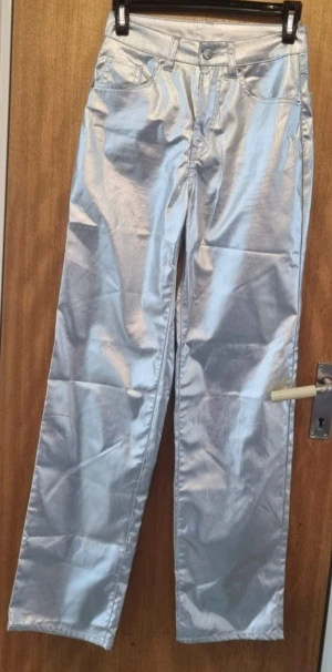Silvriga raka byxor med glans - Snygga silvriga byxor med rak passform och glansig finish. Klassisk femficksmodell med knapp och dragkedja fram. Perfekta för dig som vill sticka ut med en cool metallic-look. Materialet känns mjukt och har en lätt skimrande yta.