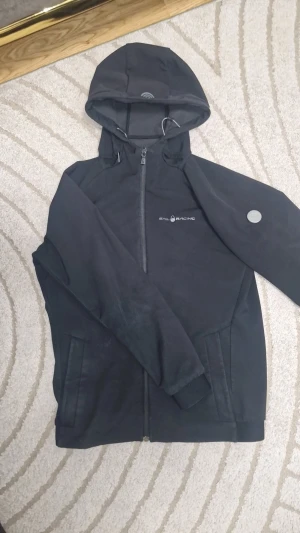 Svart vindjacka från Sail Racing - Svart vindjacka från Sail Racing med huva och dragkedja framtill. Jackan har två sidofickor, snygg logotyp på bröstet och en rund metallbadge på ärmen. Materialet är syntet och jackan har en clean, sportig look som passar perfekt till streetwear.