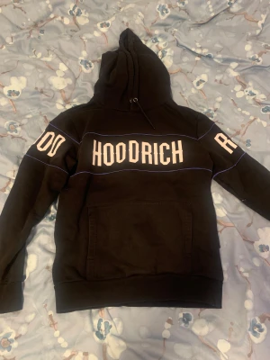 Svart och blå hoodrich hoodie - Svart hoodie från Hoodrich med stor vit logga tryckt över bröstet och ryggen. Tröjan har huva med snörning, känguruficka och blå linjer som detaljer på ärmar och över bröstet. Mjuk och skön perfekt för chill eller streetwear.