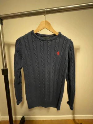 Mörkblå kabelstickad tröja  - Snygg mörkblå kabelstickad tröja från Skill med rund hals och lång ärm. Tröjan har ett rött broderat ekorrmotiv på bröstet som ger en cool detalj. Perfekt för dig som gillar klassisk stil med twist.