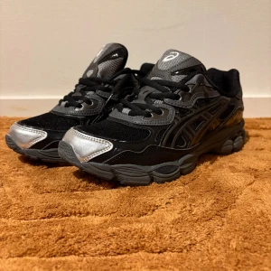 Svarta och silver Asics sneakers - Säljer ett par snygga Asics sneakers i svart och silver med coola detaljer och en unik innersula med grafiskt tryck. Skorna har snörning, platt sula och meshpartier för extra komfort. Perfekt för dig som gillar streetstyle och vill sticka ut med din look.