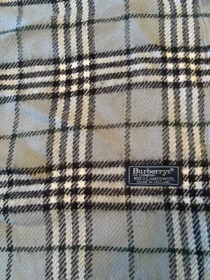 Rutig halsduk från Burberrys i ull - Snygg rutig halsduk från Burberrys i 100% lammull, tillverkad i England. Färgerna går i grått, svart och vitt med klassiskt rutmönster och tvinnade fransar i ändarna. Perfekt för dig som gillar tidlös design och vill ha något som sticker ut.