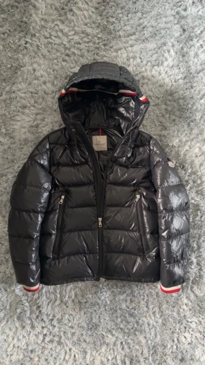 Svart Moncler pufferjacka med huva - Svart pufferjacka från Moncler med glansig finish, huva och två dragkedjeförsedda fickor. Jackan har Moncler-logga på ärmen och coola rödvita detaljer vid huva och ärmslut. Perfekt för dig som vill ha en snygg och varm jacka med streetkänsla.