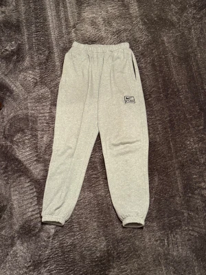 Nike x Stussy grå mjukisbyxor  - Grå mjukisbyxor från Nike x Stussy med broderad logga på benet. Byxorna har resår i midjan och vid bensluten, samt sidofickor. Tillverkade i mjuk bomullsblandning för maximal komfort och avslappnad stil. 