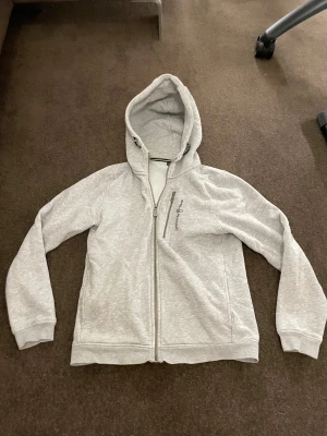 Ljusgrå hoodie från Sail Racing - Ljusgrå hoodie från Sail Racing med hel dragkedja och extra dragkedjeficka på bröstet. Tröjan har broderad logga, huva och långa ärmar. Mjuk insida i fleece och ribbade muddar vid ärmslut och nederkant. Perfekt för chill eller utomhus.