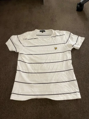 Vit randig t-shirt från Lyle & Scott vintage Stl 170 - Vit t-shirt från Lyle & Scott med svarta horisontella ränder och klassisk rund hals. På bröstet sitter den ikoniska broderade gula fågeln. T-shirten är kortärmad och gjord i mjuk bomull, perfekt för en chill och stilren look.