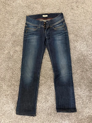 Blå straight jeans från Pepe Jeans - Snygga blå jeans från Pepe Jeans med låg midja och raka ben. Jeansen har klassisk femficksdesign, dubbla knappar i gylfen och kontrastsömmar. Bakfickorna har broderade detaljer och en läderpatch med logga. Perfekt för dig som gillar en clean och tidlös look.