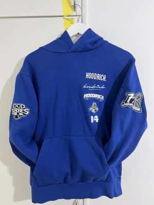 Blå Hoodie från Hoodrich med tryck - Snygg blå hoodie från Hoodrich med stora vita tryck och broderade detaljer på både fram- och baksida samt på huvan. Tröjan har känguruficka, långa ärmar och coola patches på båda armarna. Perfekt för dig som gillar streetwear och vill sticka ut.