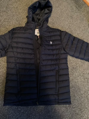 Mörkblå pufferjacka från US Polo Assn - Säljer en mörkblå pufferjacka från US Polo Assn med huva och dragkedja. Jackan har horisontella quiltade paneler, två fickor med dragkedja och diskret logga på bröstet. Perfekt för kyliga dagar och riktigt snygg till streetwear-stilen. Den är aldrig använd