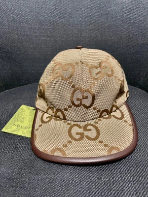 Gucci keps  - Exklusiv keps från Gucci i beige och brunt med det klassiska GG-logomönstret över hela kepsen. Kepsen har en böjd skärm med brunt kantband och justerbar rem baktill. Materialet är slitstark bomull och polyester, perfekt för dig som vill sticka ut med lyxig streetstyle.