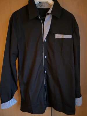 Svart skjorta från Royal med ränder - Svart skjorta från Royal med snygga randiga detaljer på insidan av kragen, knappslån, bröstfickan och ärmsluten. Skjortan har klassisk krage, knappar framtill och långärmad design. Perfekt för dig som gillar stilrena plagg med en twist.