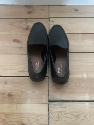 Svarta loafers från Polo Ralph Lauren - Säljer ett par stilrena svarta loafers från Polo Ralph Lauren i storlek 42. Skorna har en klassisk rund tå, platt sula med gummidetaljer och är tillverkade i skinn med brunt innermaterial. Perfekta för dig som gillar en clean och tidlös look.