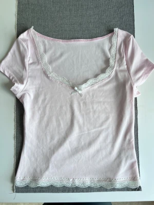 Ljusrosa topp med spets och rosett, storlek S - Ljusrosa topp med korta ärmar, v-ringad hals och vita spetskanter. Framtill sitter en liten vit rosett 🎀🤍 Materialet är mjukt och väldigt stretchigt. - Köper du innan kl 16:00 kan jag posta samma dag!