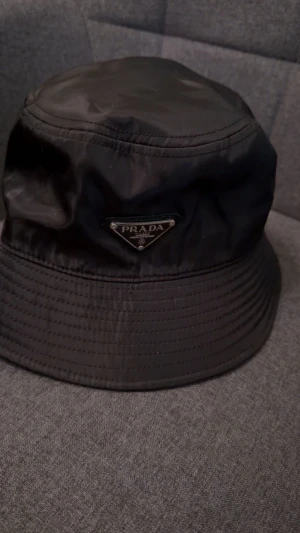 Svart bucket hat från Prada - Säljer en svart bucket hat från Prada med klassisk trekantig logga framtill. Hatten har en stilren design med bred kant och är tillverkad i glansig syntet. Perfekt för dig som vill ha en lyxig och trendig accessoar med streetkänsla.