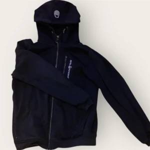 Svart hoodie från Sail Racing 160 - Svart hoodie från Sail Racing med dragkedja framtill och logga på bröstet. Tröjan har huva och är tillverkad i en mjuk bomulls- och polyestermix. Perfekt för dig som gillar sportig och clean stil.