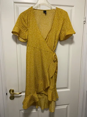 Gul omlottklänning med prickar H&M - Helt ny och oanvänd mörkgul omlottklänning från Veromoda med vita prickar, volangdetaljer och knyt i midjan. Klänningen har korta, fladdriga ärmar och v-ringning. Fint fall i tyget och knytband i sidan. 