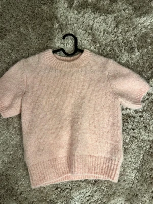 Ljusrosa stickad tröja med kort ärm - Supermjuk och fluffig ljusrosa stickad tröja med korta ärmar och ribbade muddar vid hals, ärmslut och nederkant. Perfekt för lager-på-lager och ger en söt vibe till din outfit.