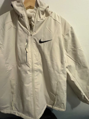Beige kofta från Nike med huva - Snygg beige kofta från Nike med svart logga på bröstet. Jackan har huva, dragkedja framtill och flera praktiska fickor med dragkedja. Perfekt för blåsiga dagar och enkel att matcha med olika outfits.