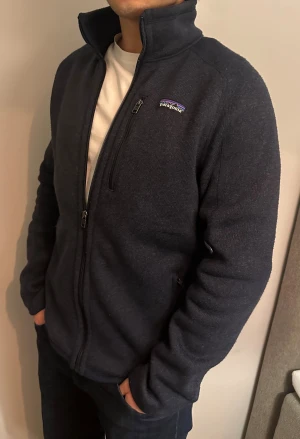 Mörkblå fleecejacka från Patagonia - Mjuk och varm mörkblå fleecejacka från Patagonia med hög krage och hel dragkedja. Jackan har en bröstficka med dragkedja och diskret logga på vänster sida. Perfekt för kyliga dagar och chill stunder. Snygg, enkel och riktigt skön passform.