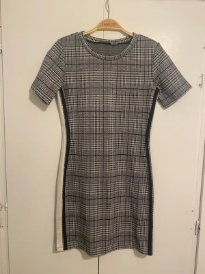 Rutig kort klänning  - Kortärmad, figurnära klänning med svartvitt rutigt mönster och kontrasterande svarta och vita ränder längs sidorna. Klassisk rund halsringning och kort längd, perfekt för en trendig look. Materialet är mjukt och stretchigt för extra komfort.