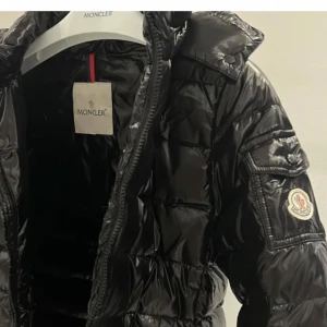 Svart Moncler pufferjacka med huva - Säljer en ÄKTA svart, glansig pufferjacka från Moncler med huva och dragkedja framtill. Jackan har Moncler-logga på ärmen och flera praktiska fickor. endast använd en vinter men en knapp på luvan krånglar men nyper man med tång är det inga problem. Inget som är sönder eller defekt. Har påse samt lappen från köpet och qr koden i visar att den är äkta:) Skriv för fler bilder och bevis! 