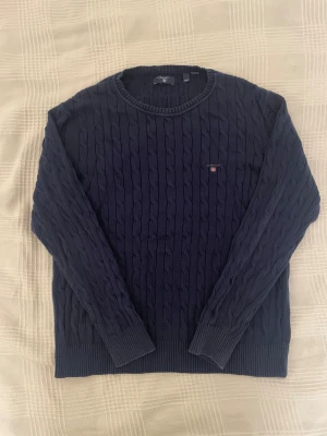 Marinblå kabelstickad tröja GANT - Snygg marinblå kabelstickad tröja från GANT med rund hals och diskret logga broderad på bröstet. Tröjan har ribbade muddar vid ärmslut och nederkant, och är gjord i mjuk bomull. Perfekt för dig som gillar klassisk stil med en modern touch.