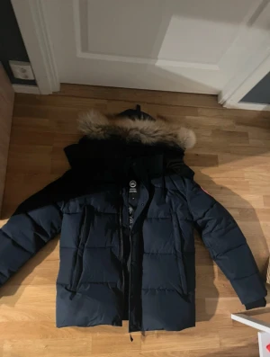 Mörkblå Canada Goose dunjacka med päls - Säljer en mörkblå dunjacka från Canada Goose med fet päls på huvan. Jackan har lång ärm, dragkedja och knappar framtill, samt klassisk logga på ärmen. Insidan är fodrad med glansigt material och jackan är riktigt varm och skön för kalla dagar.