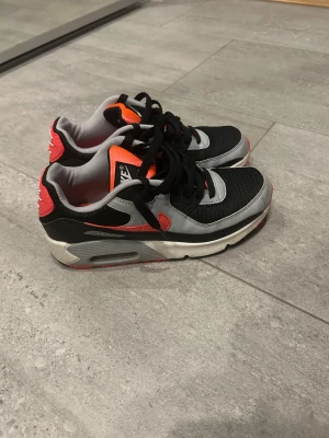 Nike Air Max 90 svart/röd/grå sneakers - Nike Air Max 90 sneakers med svart mesh, grå och röda detaljer samt vit sula. Klassisk Air Max-dämpning, svart snörning och rund tå. Snygg kontrast mellan färgerna och ikonisk design med synlig luftbubbla i sulan. Perfekt för dig som gillar streetstyle och sportig vibe. Tvättar de innan jag skickar pris kan diskuteras 