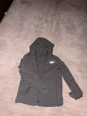 Nike grå vindjacka Dri-FIT M - Nike Performance FORM vindjacka i smoke grey med reflekterande silvrig logga på bröstet. Jackan har huva med snörning, hel dragkedja och är tillverkad i lätt, syntetiskt Dri-FIT-material som andas. Perfekt för träning och aktiva dagar.