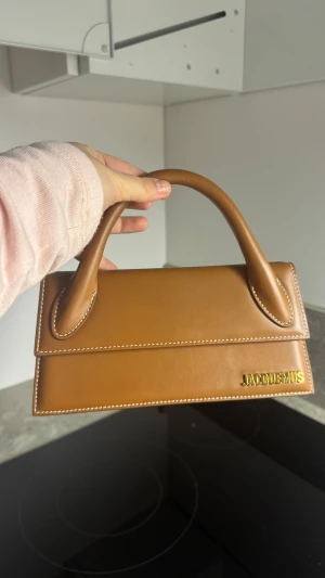 Brun Jacquemus handväska  - Säljer en brun handväska från Jacquemus i slätt skinn med vit kontrastsöm och guldfärgad logga framtill. Väskan har en stilren, rektangulär form och ett stadigt handtag. Perfekt för dig som gillar minimalistisk och trendig design.