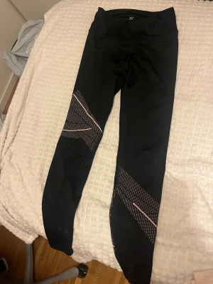 Svarta leggings med rosa detaljer - Säljer ett par svarta leggings från H&M med snygga rosa och vita mönsterdetaljer på benen. De har en tight passform och är gjorda i stretchigt syntetmaterial, perfekta för träning eller chill. Midjan är normal och designen är modern och sportig.