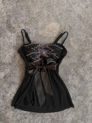 Svart blommig linnetopp med rosett - Supersnygg svart linnetopp med tunna axelband och blommigt broderi i silvertråd över bysten. Dekorerad med pärlor och en stor rosett i midjan som ger extra stilpoäng. Toppen är lätt utsvängd nedtill och har en feminin passform, perfekt för att sticka ut.