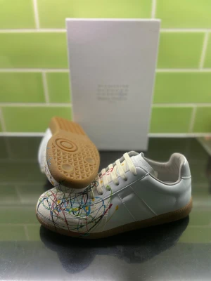 Maison Margiela sneakers med färgstänk - Unika vita sneakers från Maison Margiela med färgstänk i blått, rött, gult, grönt och svart. Skorna har beige snörning, gummisula och är tillverkade i skinn och mocka. Perfekt för dig som vill sticka ut med en kreativ och konstnärlig vibe.