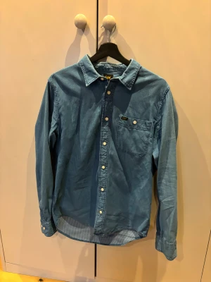Blå manchesterskjorta från Lee M - Snygg blå skjorta från Lee i mjuk manchester, regular fit. Skjortan har klassisk krage, vita knappar och en bröstficka med Lee-logga. Perfekt för dig som gillar avslappnad stil med lite retrovibe. Materialet är 100% bomull och den har långa ärmar.