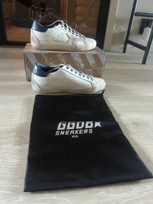 Golden Goose vita sneakers med blå detalj - Golden Goose sneakers i vitt skinn med blå hälkappa och grå mockadetaljer. Klassisk stjärnlogga på sidan och GGDB/SSTAR-tryck. Platt sula och snörning framtill. Snyggt slitna detaljer för en cool look. Kommer med originalkartong och dustbag.
