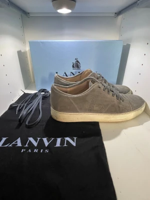 Grå mockasneakers från Lanvin - Snygga grå sneakers från Lanvin i mjuk mocka med vit platt sula och rund tå. Skorna har klassisk snörning och diskret perforerat L på sidan. Innersulan är i beige skinn och de kommer med extra skosnören och originalpåse i och låda. TAR BYTEN‼️