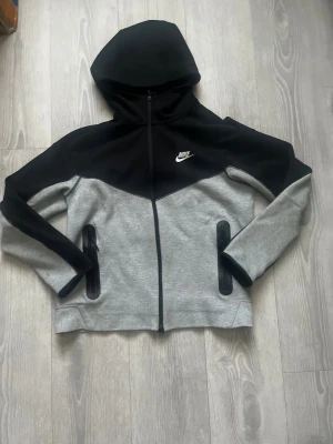 Nike hoodie svart och grå med dragkedja - Nike hoodie i svart och grått med hel dragkedja framtill och två fickor med dragkedja. Snyggt broderad Nike-logga på bröstet. Tröjan har huva och är långärmad, perfekt för chill eller träning. Materialet är mjukt och känns som bomull och polyester.