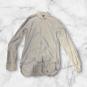 ljus blå långärmad skjorta Polo Ralph Lauren - Snygg beige skjorta från Polo Ralph Lauren med klassisk krage och diskret logga på bröstet. Skjortan har långa ärmar, knappar framtill och är tillverkad i mjuk bomull. Perfekt för dig som gillar stilrena och tidlösa plagg. Slim fit och passar M/L