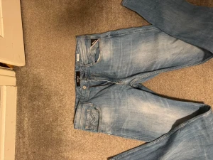 Replay ljusblå jeans med raka ben - Säljer ett par ljusblå jeans från Replay med klassisk femficksdesign och raka ben. Jeansen har snygga slitningar och detaljer som Replay-logga på knapp och etikett. Materialet är mjukt denim och de har dragkedja i gylfen.