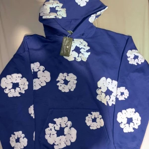 Blå hoodie från Denim Tears - Säljer en blå hoodie från Denim Tears med vita blommönster över hela plagget. Hoodien har en klassisk design med en stor ficka framtill och en justerbar huva. Perfekt för en avslappnad stil.