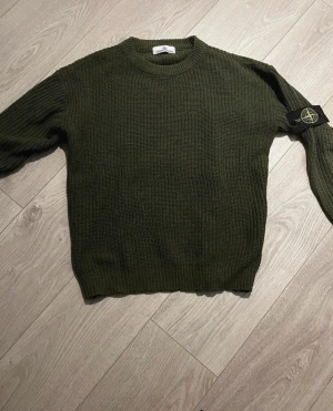 Grön stickad tröja från Stone Island - Säljer en mörkgrön stickad tröja från Stone Island med klassisk rund hals och ribbade muddar. Tröjan har lång ärm och den ikoniska Stone Island-patchen på vänster arm. Perfekt för dig som gillar streetwear och snygga detaljer. Säljer då den var lite för stor för mig! Kom med prisförslag!