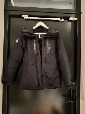 Svart Sail Racing dunjacka Gore-Tex S - Svart dunjacka från Sail Racing med Gore-Tex-material, perfekt för kalla dagar. Jackan har två vertikala bröstfickor med dragkedja, hög krage och logotyp på ärmen.