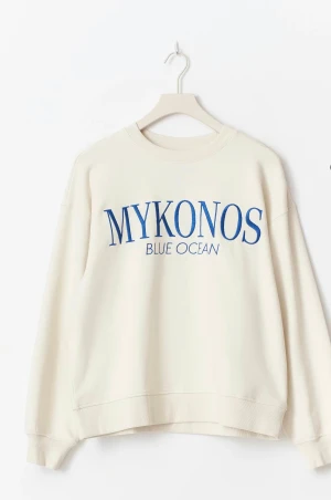 Vit sweatshirt med blå text MYKONOS - Säljer en vit sweatshirt med trycket MYKONOS BLUE OCEAN i blått framtill. Tröjan har rund hals, ribbade muddar och lång ärm. Perfekt för dig som gillar clean och chill stil. Materialet är mjuk bomull och passformen är normal.
