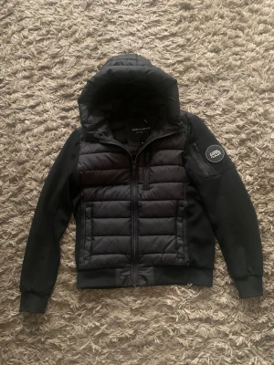 Svart pufferjacka Supply & Demand M - Svart pufferjacka från Supply & Demand med huva och quiltad framsida. Jackan har dragkedja, ribbade muddar och en patch med logga på ärmen. storlek M men passar också S. Jackan är i väldigt bra skick, hör av dig vid frågor! (pris kan diskuteras)