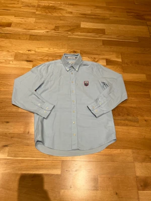 Ljusblå GANT Le Mans skjorta - Snygg ljusblå skjorta från GANT x Le Mans med broderad patch på bröstet. Klassisk button-down krage, vita knappar och lång ärm. Tillverkad i bomull och perfekt för en clean och stilren look. (Ai beskrivning så hör av dig om du har några frågor). Priset är prutbart vid enkel affär