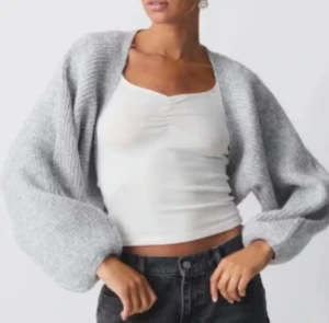 Grå stickad bolerotopp - Säljer en grå stickad bolerotopp med breda, lösa ärmar och ribbad struktur. Perfekt att slänga över en linne eller topp för en chill och trendig look. Toppens material är mjukt och ger en cozy vibe, samtidigt som den är lätt att styla till jeans eller kjol. Helt ny! Slut online!