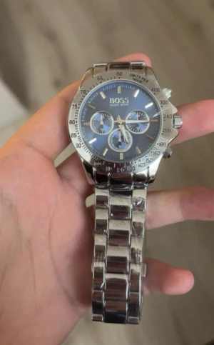 Hugo Boss silverfärgad klocka - Stilren kronografklocka från Hugo Boss med blå urtavla och tre mindre visartavlor. Klockan har en robust länkad armlänk i blankt silverfärgat stål och tydliga markeringar på boetten. Perfekt accessoar för dig som gillar klassisk design med modern touch.